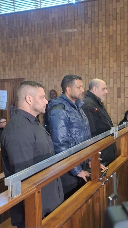 Three men stand in a courtroom, appearing serious and attentive. They are behind a wooden railing against a backdrop of brick walls, suggesting a formal legal setting.