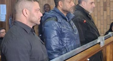 Three men stand in a courtroom, appearing serious and attentive. They are behind a wooden railing against a backdrop of brick walls, suggesting a formal legal setting.