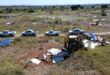 Aerial view of a grassy field with scattered solar panels and debris. A bulldozer demolishes structures as white trucks are lined up nearby, conveying a scene of deconstruction.