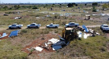 Aerial view of a grassy field with scattered solar panels and debris. A bulldozer demolishes structures as white trucks are lined up nearby, conveying a scene of deconstruction.
