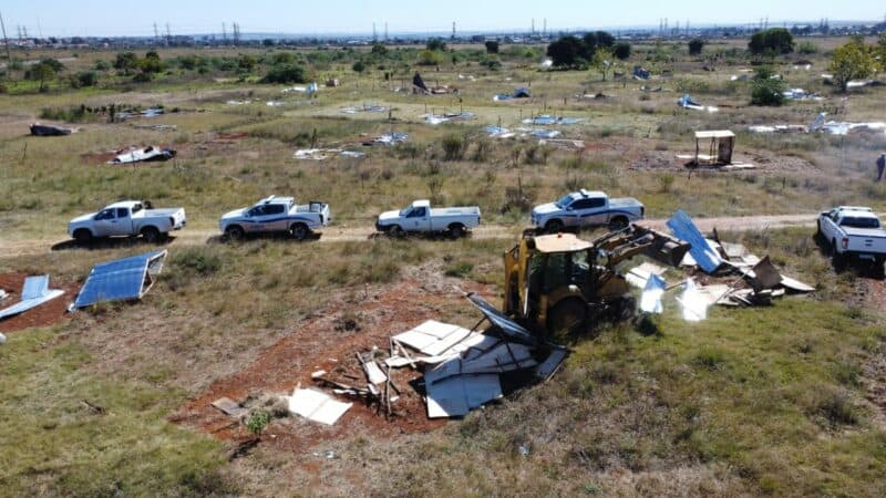 Aerial view of a grassy field with scattered solar panels and debris. A bulldozer demolishes structures as white trucks are lined up nearby, conveying a scene of deconstruction.