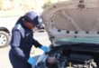 A police officer inspects a car engine, wearing a blue uniform and gloves, with the hood open. The scene is outdoors on a sunny day.