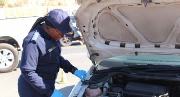 A police officer inspects a car engine, wearing a blue uniform and gloves, with the hood open. The scene is outdoors on a sunny day.