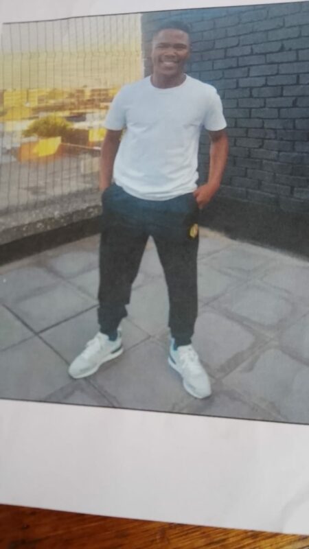 Young man smiling in a white t-shirt and dark pants, standing on a tiled terrace with a brick wall and city view in the background; casual and relaxed mood.