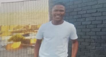 Young man smiling in a white t-shirt and dark pants, standing on a tiled terrace with a brick wall and city view in the background; casual and relaxed mood.
