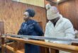 A woman in a blue outfit and a man in a white coat and knit hat stand at a wooden courtroom bench, appearing serious and focused.