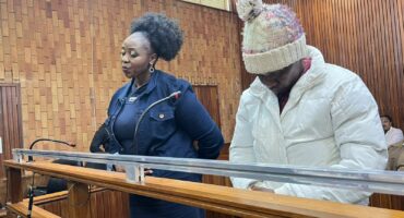 A woman in a blue outfit and a man in a white coat and knit hat stand at a wooden courtroom bench, appearing serious and focused.