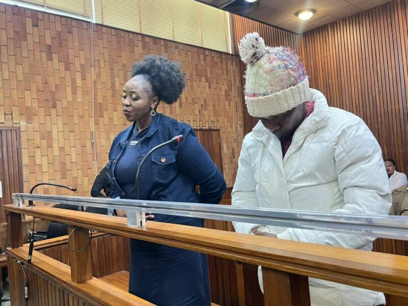 A woman in a blue outfit and a man in a white coat and knit hat stand at a wooden courtroom bench, appearing serious and focused.