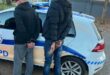 Two men stand handcuffed behind their backs next to a police car on a residential street. Their faces are obscured.