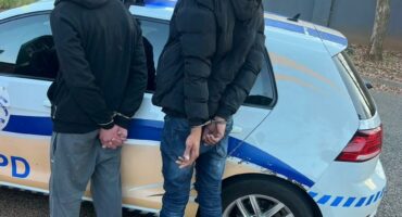 Two men stand handcuffed behind their backs next to a police car on a residential street. Their faces are obscured.
