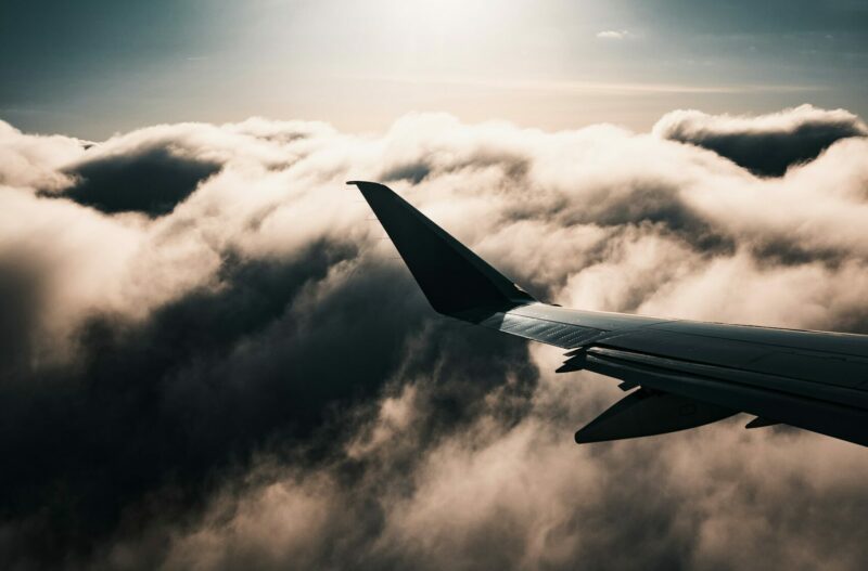 Airplane wing above dramatic, cloudy sky with sun setting, casting a serene, warm glow through the clouds. The mood is peaceful and contemplative.