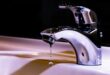 A shiny chrome faucet over a white sink drips water, captured in mid-air against a dark background, conveying a sense of stillness and precision.