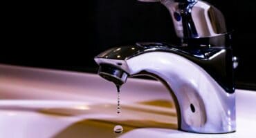 A shiny chrome faucet over a white sink drips water, captured in mid-air against a dark background, conveying a sense of stillness and precision.
