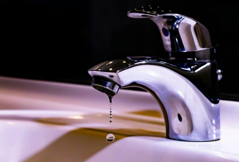 A shiny chrome faucet over a white sink drips water, captured in mid-air against a dark background, conveying a sense of stillness and precision.