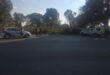 Two vehicles, one marked "R&D RISK" and one police car, are parked on either side of a road with police tape and several people standing nearby. Trees and bushes are in the background.