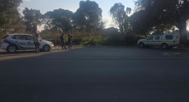 Two vehicles, one marked "R&D RISK" and one police car, are parked on either side of a road with police tape and several people standing nearby. Trees and bushes are in the background.
