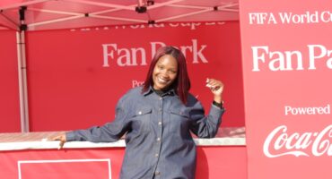 A smiling woman poses at a FIFA World Cup Fan Park event, showcasing a vibrant red backdrop with Coca-Cola branding.