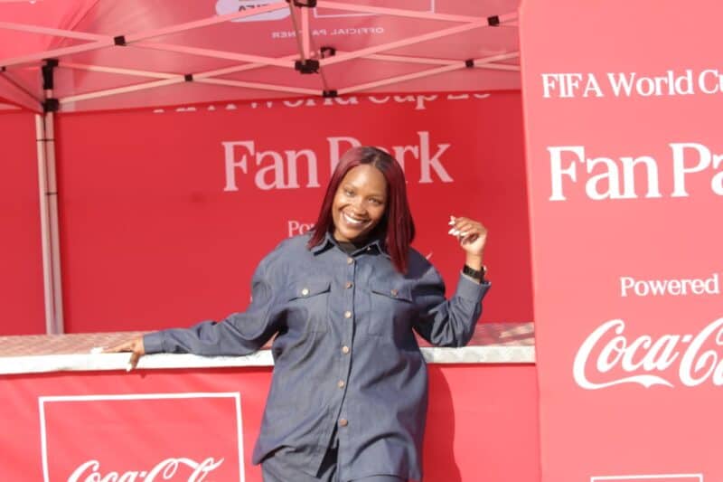 A smiling woman poses at a FIFA World Cup Fan Park event, showcasing a vibrant red backdrop with Coca-Cola branding.