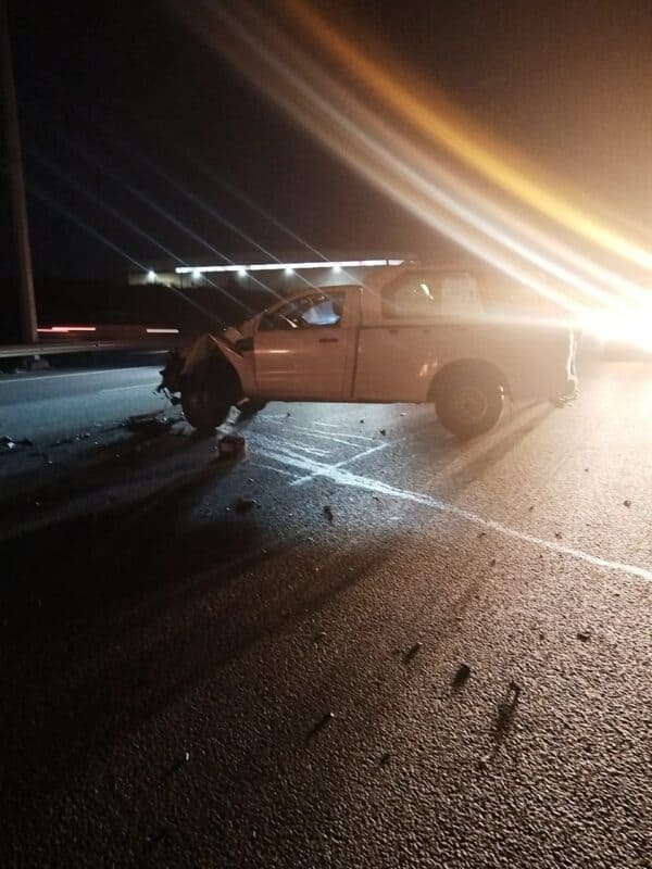 White pickup truck with front-end damage stopped on a dark road at night with debris scattered around and bright headlights in the background.