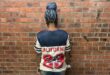 Person wearing a California jersey with number 23, standing against a brick wall, showcasing braided hairstyle from the back.