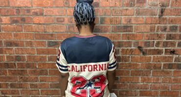 Person wearing a California jersey with number 23, standing against a brick wall, showcasing braided hairstyle from the back.
