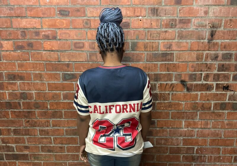Person wearing a California jersey with number 23, standing against a brick wall, showcasing braided hairstyle from the back.