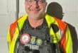A smiling man in a safety vest and tactical gear stands against a wall, showcasing various badges and patches on his uniform.
