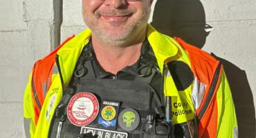 A smiling man in a safety vest and tactical gear stands against a wall, showcasing various badges and patches on his uniform.