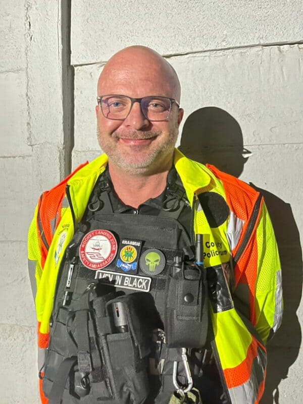 A smiling man in a safety vest and tactical gear stands against a wall, showcasing various badges and patches on his uniform.