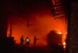 Firefighters battling a large blaze in a warehouse, with intense flames and smoke filling the scene during nighttime operations.