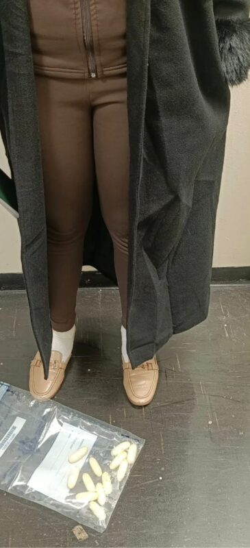 A person wearing a black coat and brown leggings stands over a clear bag containing white capsules on a dark floor.