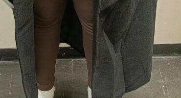 A person wearing a black coat and brown leggings stands over a clear bag containing white capsules on a dark floor.