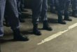 Row of uniformed individuals standing in black boots on a concrete floor, with visible gray pants and a white line marking the ground.