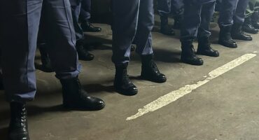 Row of uniformed individuals standing in black boots on a concrete floor, with visible gray pants and a white line marking the ground.