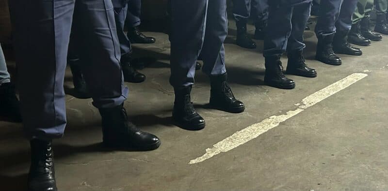 Row of uniformed individuals standing in black boots on a concrete floor, with visible gray pants and a white line marking the ground.