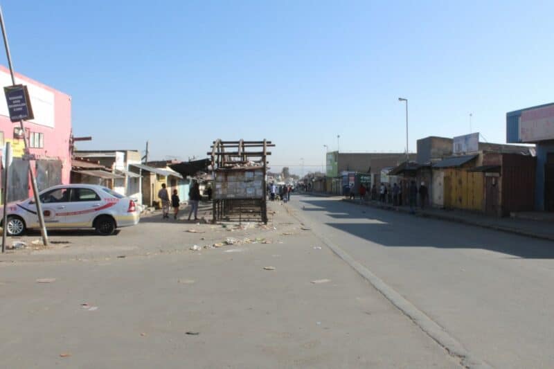 A street scene with people walking past buildings and market stalls under a clear blue sky. Some structures are weathered and colorful.