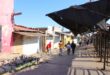 A street scene with people walking past buildings and market stalls under a clear blue sky. Some structures are weathered and colorful.