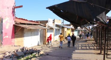A street scene with people walking past buildings and market stalls under a clear blue sky. Some structures are weathered and colorful.