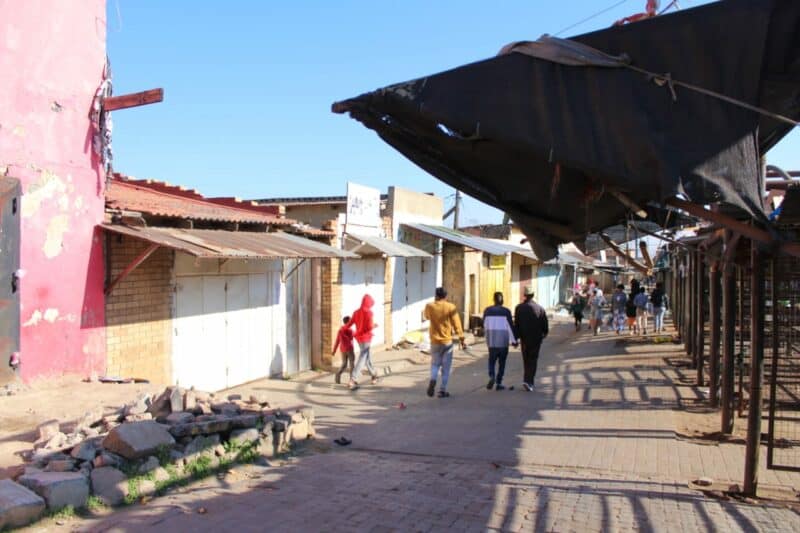 A street scene with people walking past buildings and market stalls under a clear blue sky. Some structures are weathered and colorful.