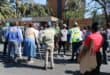 A group of people gathered outside the Kempton Park SAPS building, some holding signs, under a clear blue sky.
