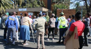 A group of people gathered outside the Kempton Park SAPS building, some holding signs, under a clear blue sky.