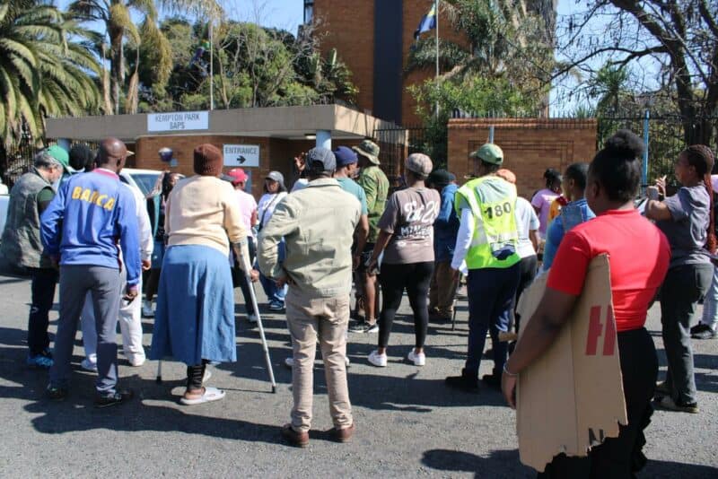 A group of people gathered outside the Kempton Park SAPS building, some holding signs, under a clear blue sky.