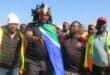 A group of men, some in safety vests, gather outdoors. One man holds a South African flag, while others look on attentively.
