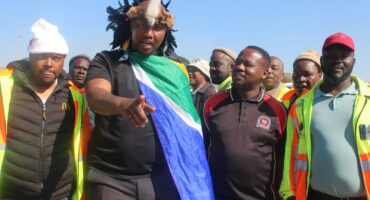 A group of men, some in safety vests, gather outdoors. One man holds a South African flag, while others look on attentively.