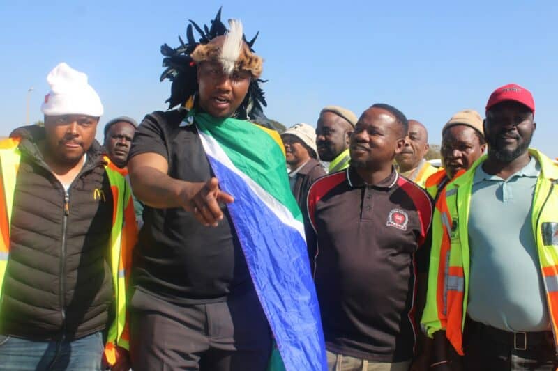 A group of men, some in safety vests, gather outdoors. One man holds a South African flag, while others look on attentively.