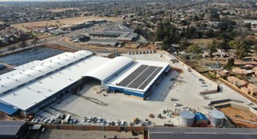 An aerial view shows a large industrial complex with white roofs, solar panels, a paved parking lot, and two large cylindrical water tanks nearby.