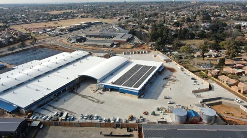 An aerial view shows a large industrial complex with white roofs, solar panels, a paved parking lot, and two large cylindrical water tanks nearby.