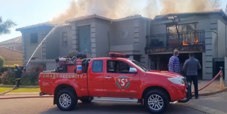 A red security truck parked in front of a house with smoke and flames visible from the roof, while firefighters work to extinguish the fire.