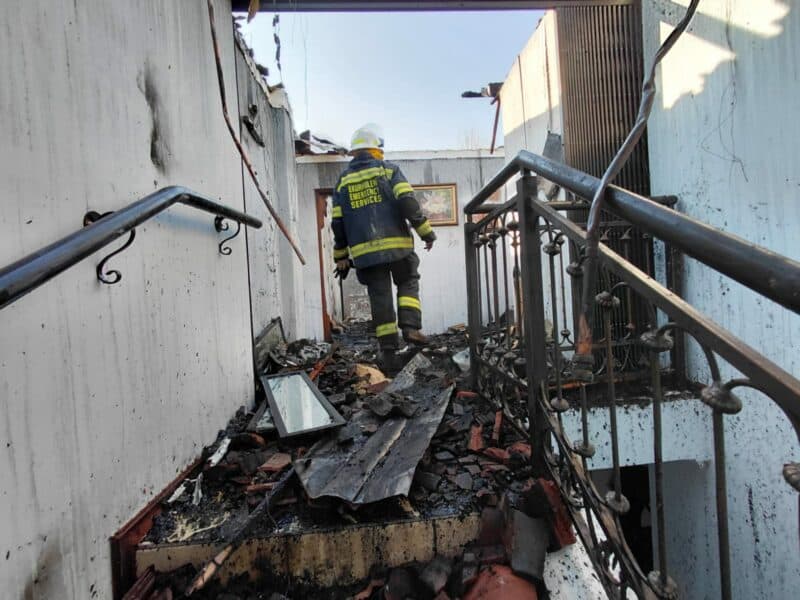 A firefighter in uniform inspects a damaged interior with debris and charred materials after a fire incident.