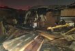 Damaged structure with burnt wooden beams and debris scattered on the ground, indicating a recent fire incident at night.